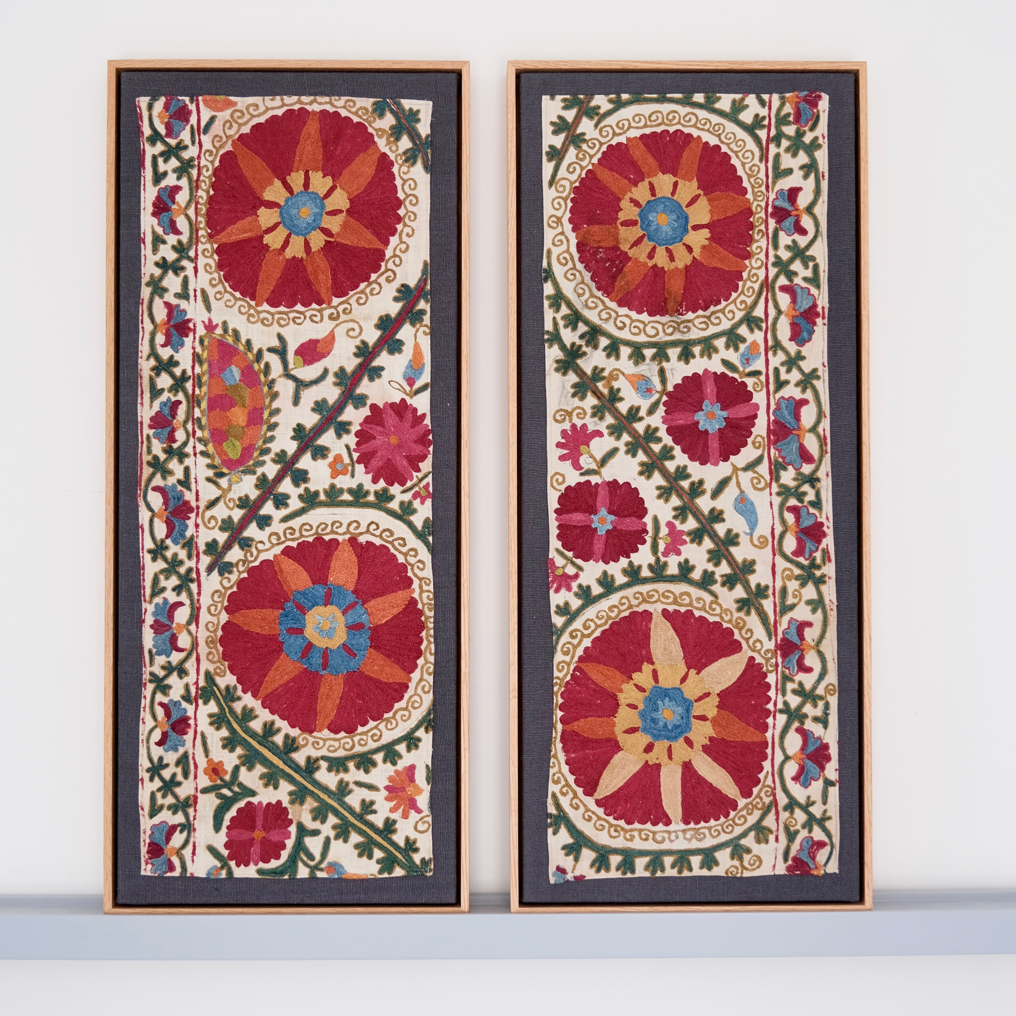 Two framed antique suzanis, embroidered textile artworks with floral patterns on a white background