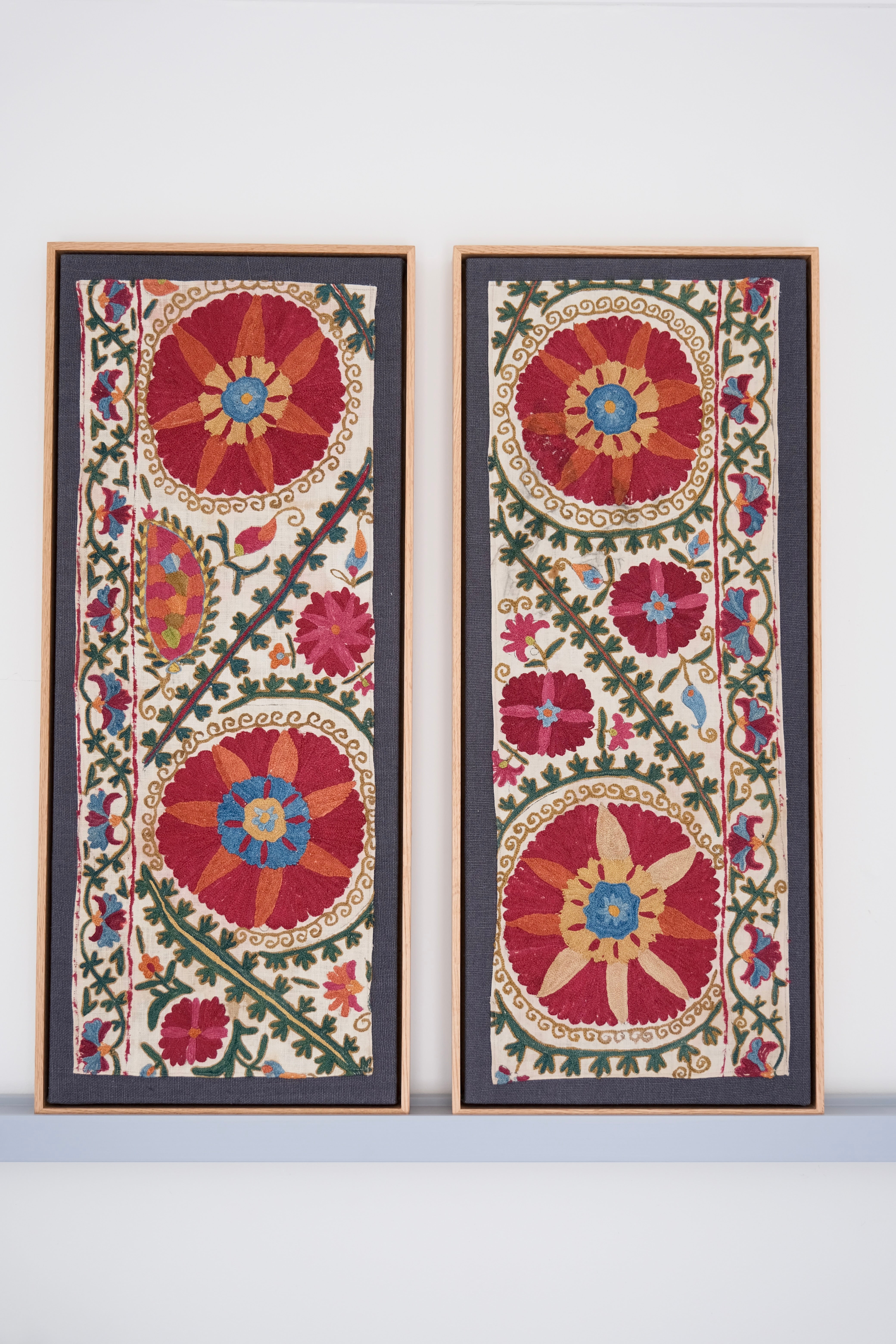 Two framed antique suzanis, embroidered textile artworks with floral patterns on a white background