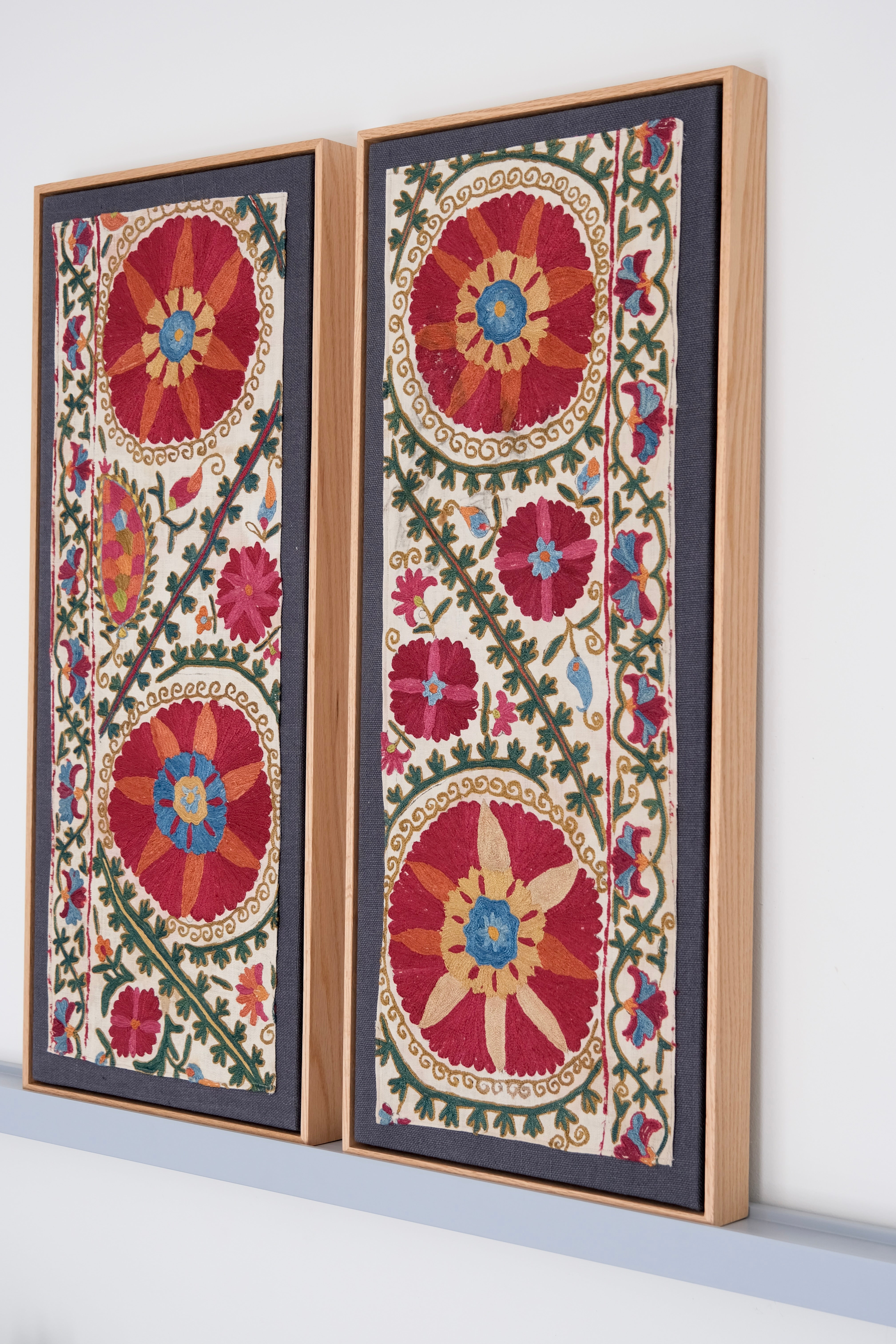 Two framed antique suzanis, embroidered textile artworks with floral patterns on a white background