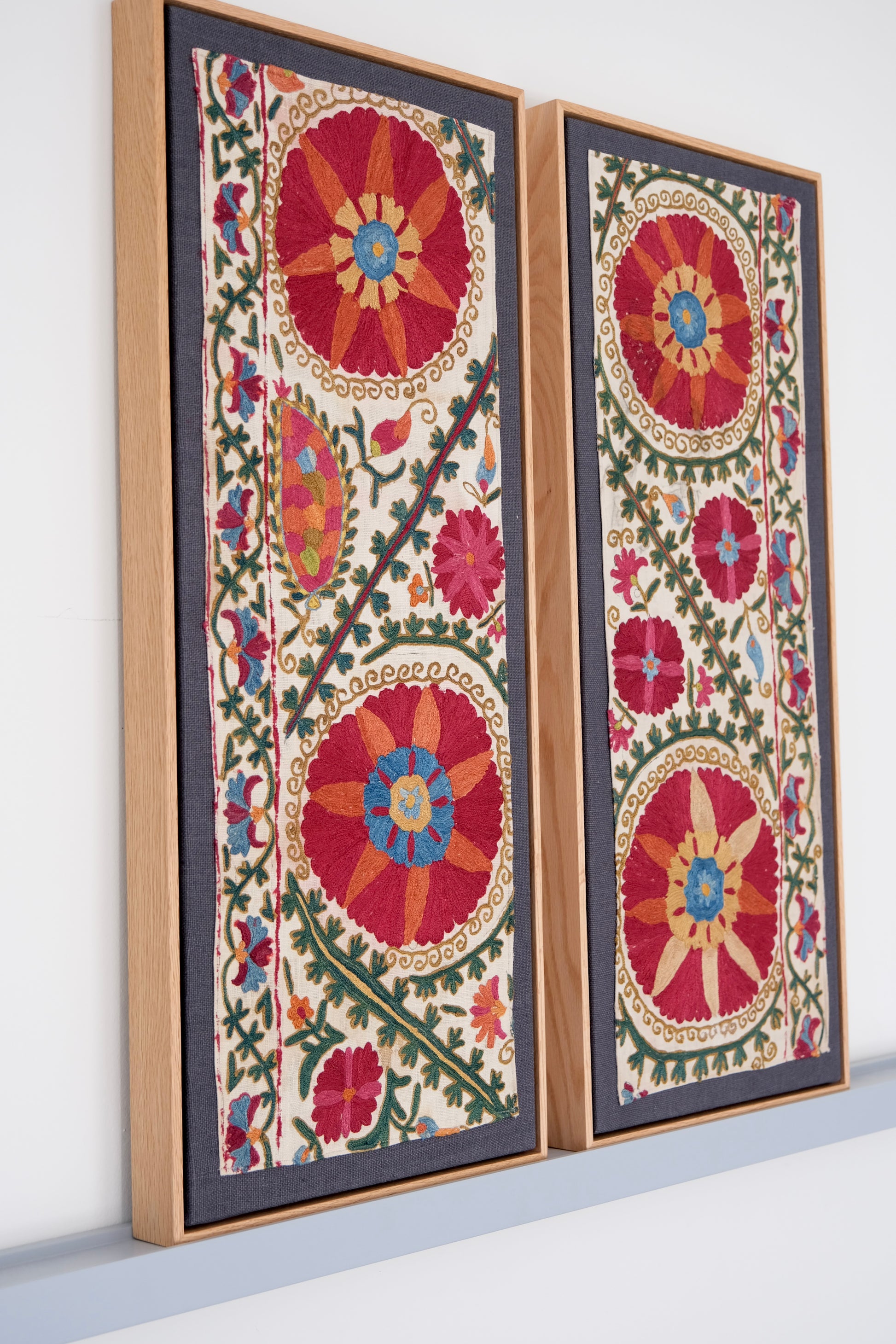 Two framed antique suzanis, embroidered textile artworks with floral patterns on a white background