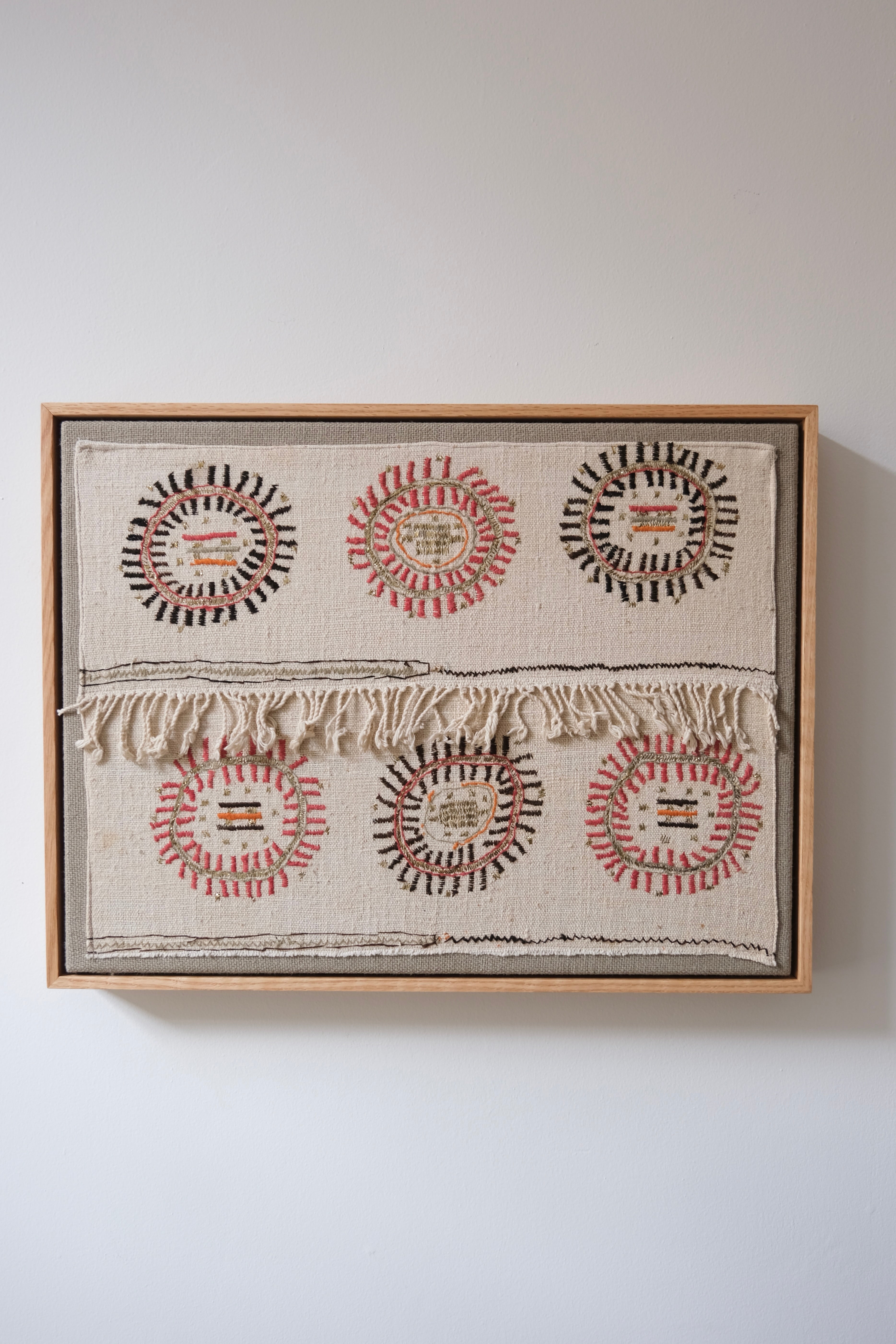 A framed textile wall hanging with six circular woven motifs in black, red, and beige on a cream fabric background, suspended against a plain light gray wall.
