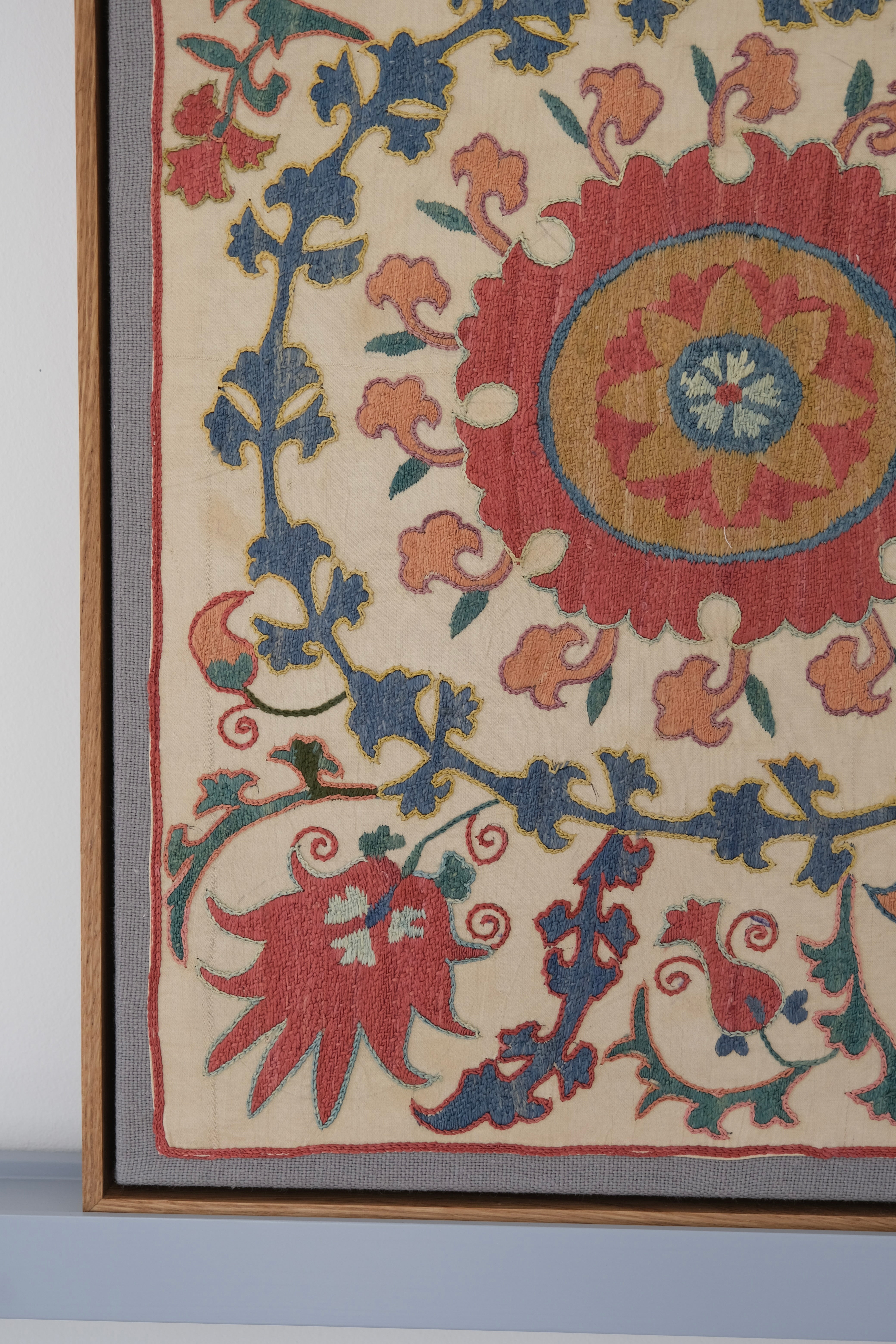 Framed suzani, embroidered textile art, wooden frame, white with pink and blue design, close-up