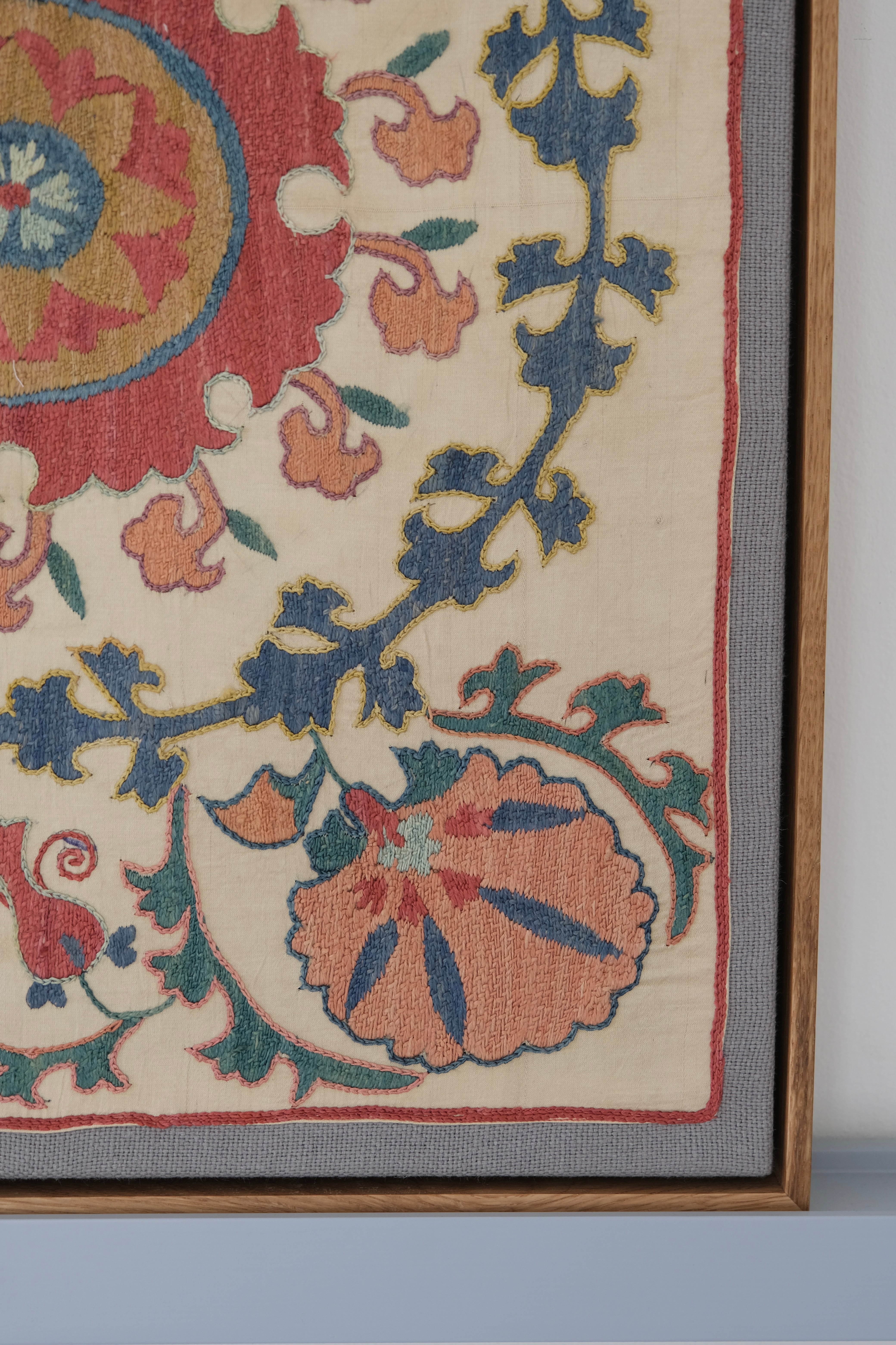 Framed suzani, embroidered textile art, wooden frame, white with pink and blue design, close-up