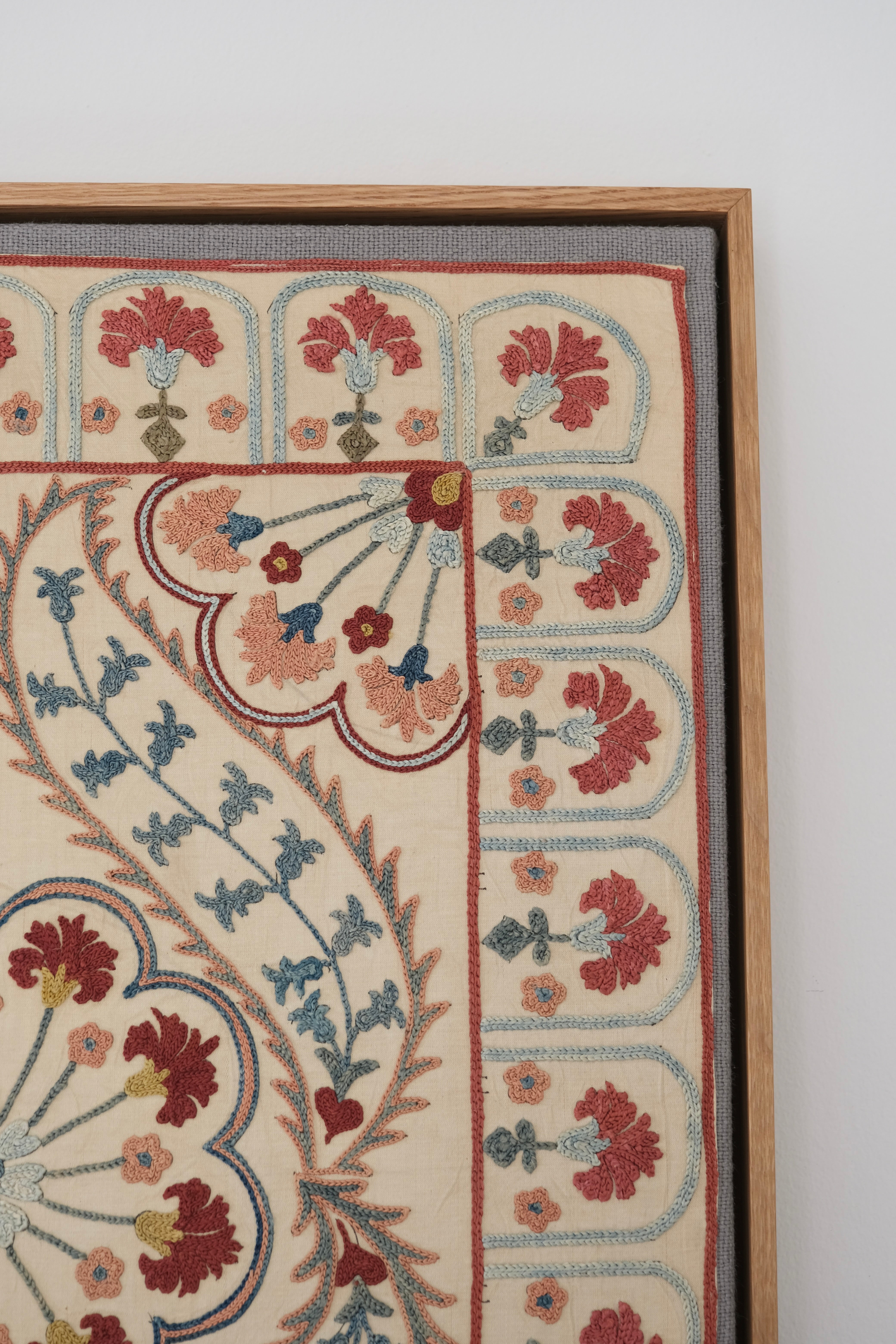 Framed suzani, embroidered textile art, wooden frame, white with blue and pink design, close-up