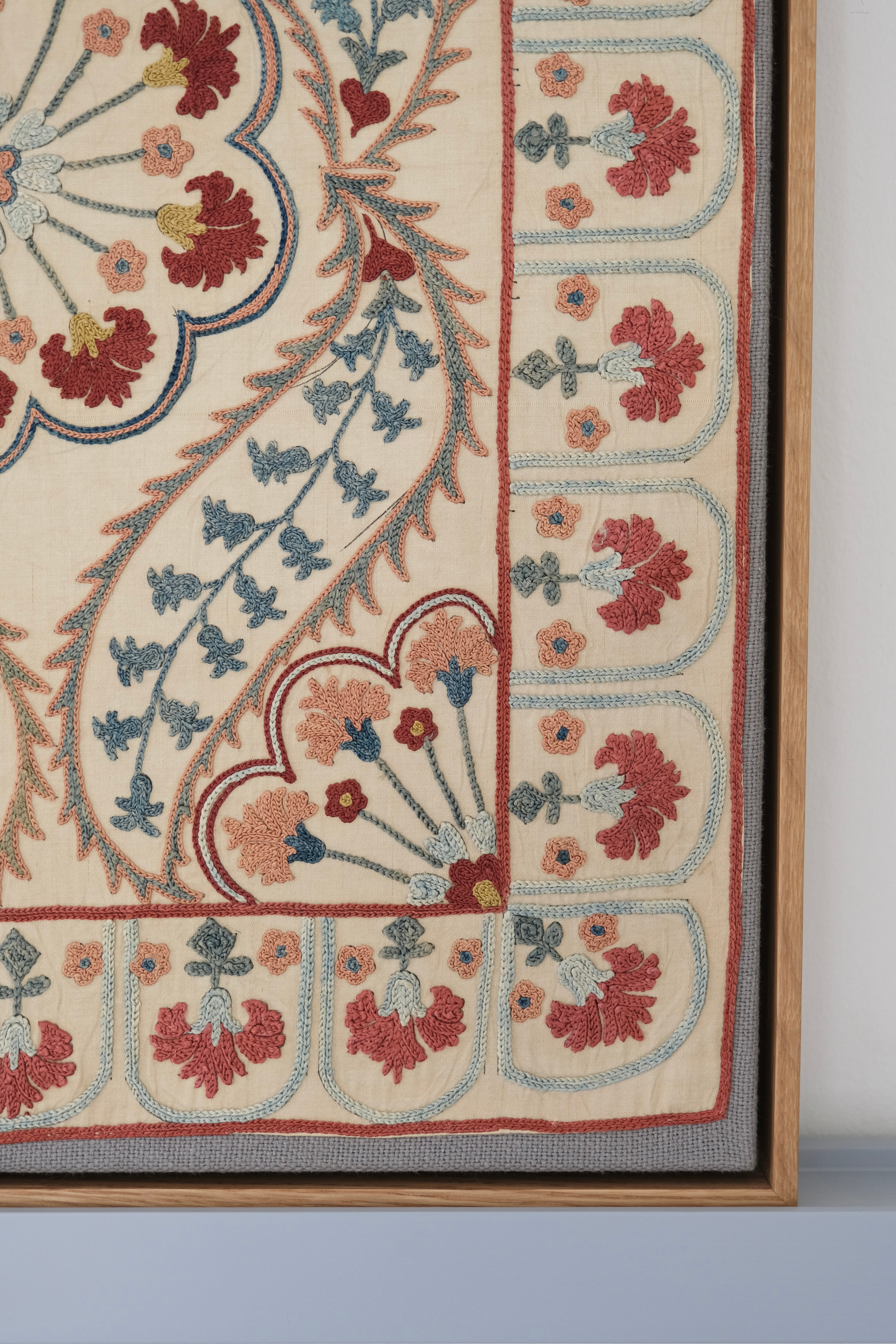 Framed suzani, embroidered textile art, wooden frame, white with blue and pink design, close-up