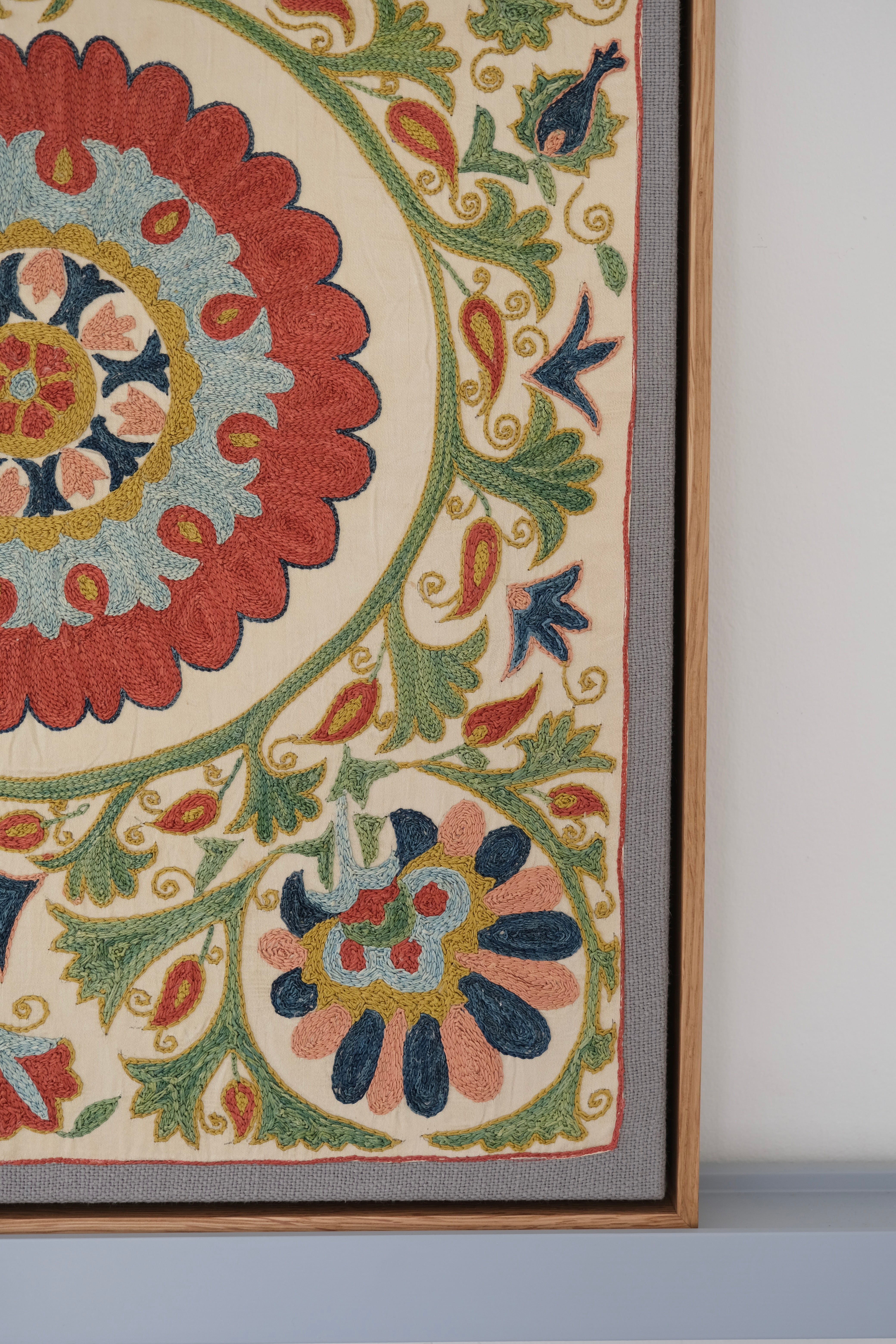 Framed suzani, embroidered textile art, wooden frame, white with green, blue and red floral design, close-up