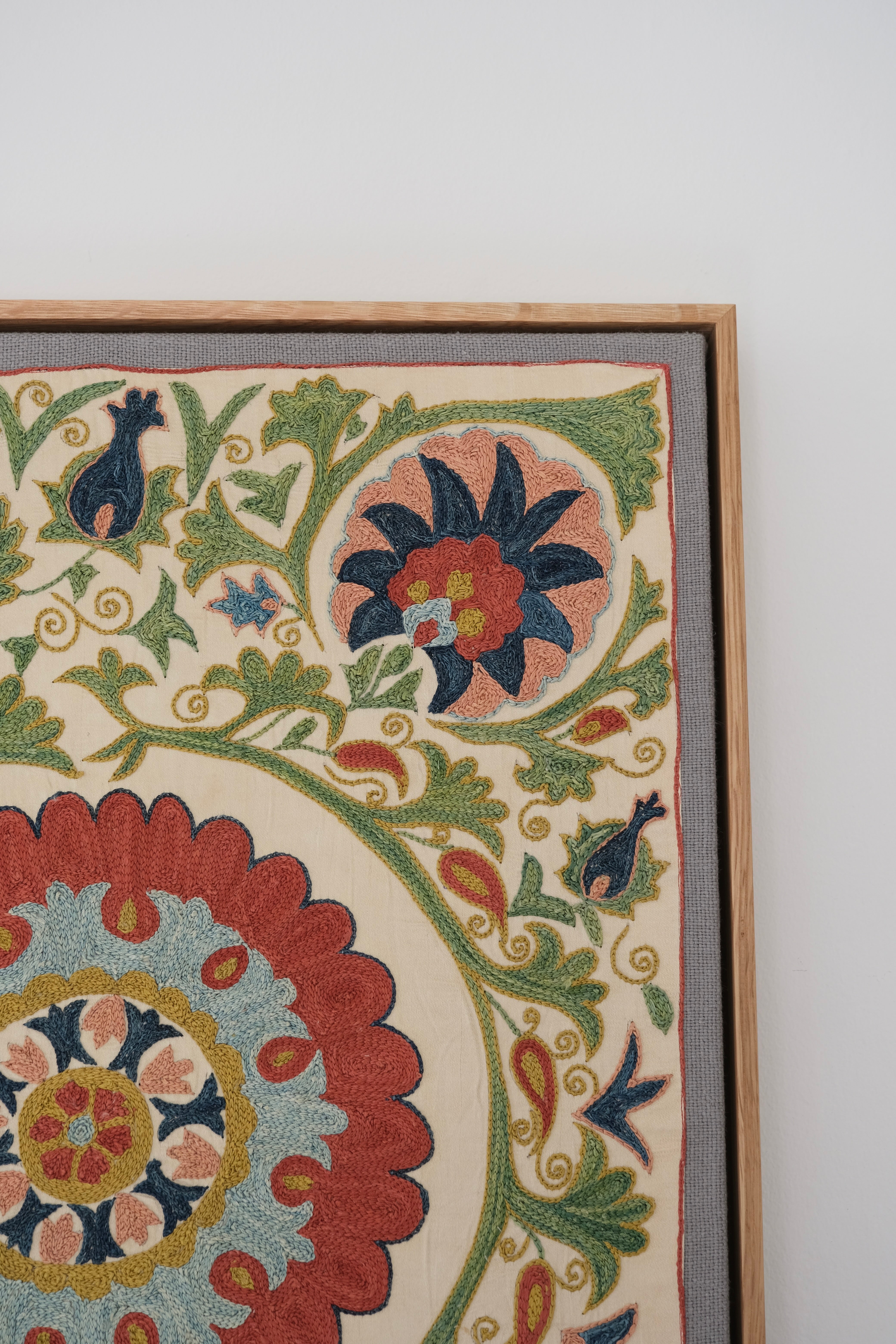 Framed suzani, embroidered textile art, wooden frame, white with green, blue and red floral design, close-up