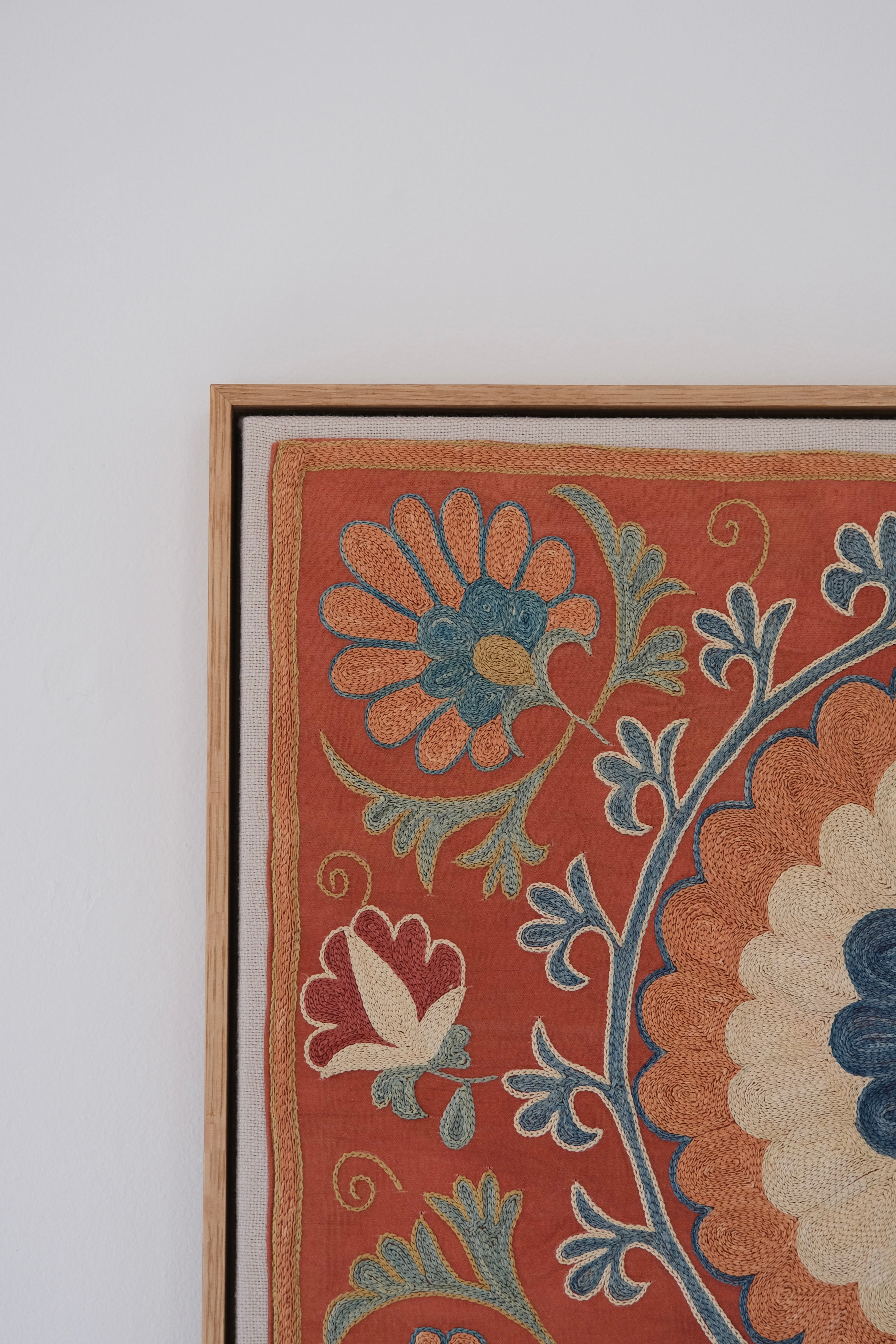 Framed suzani, embroidered textile art, wooden frame, dark orange with blue and green floral motifs, close-up