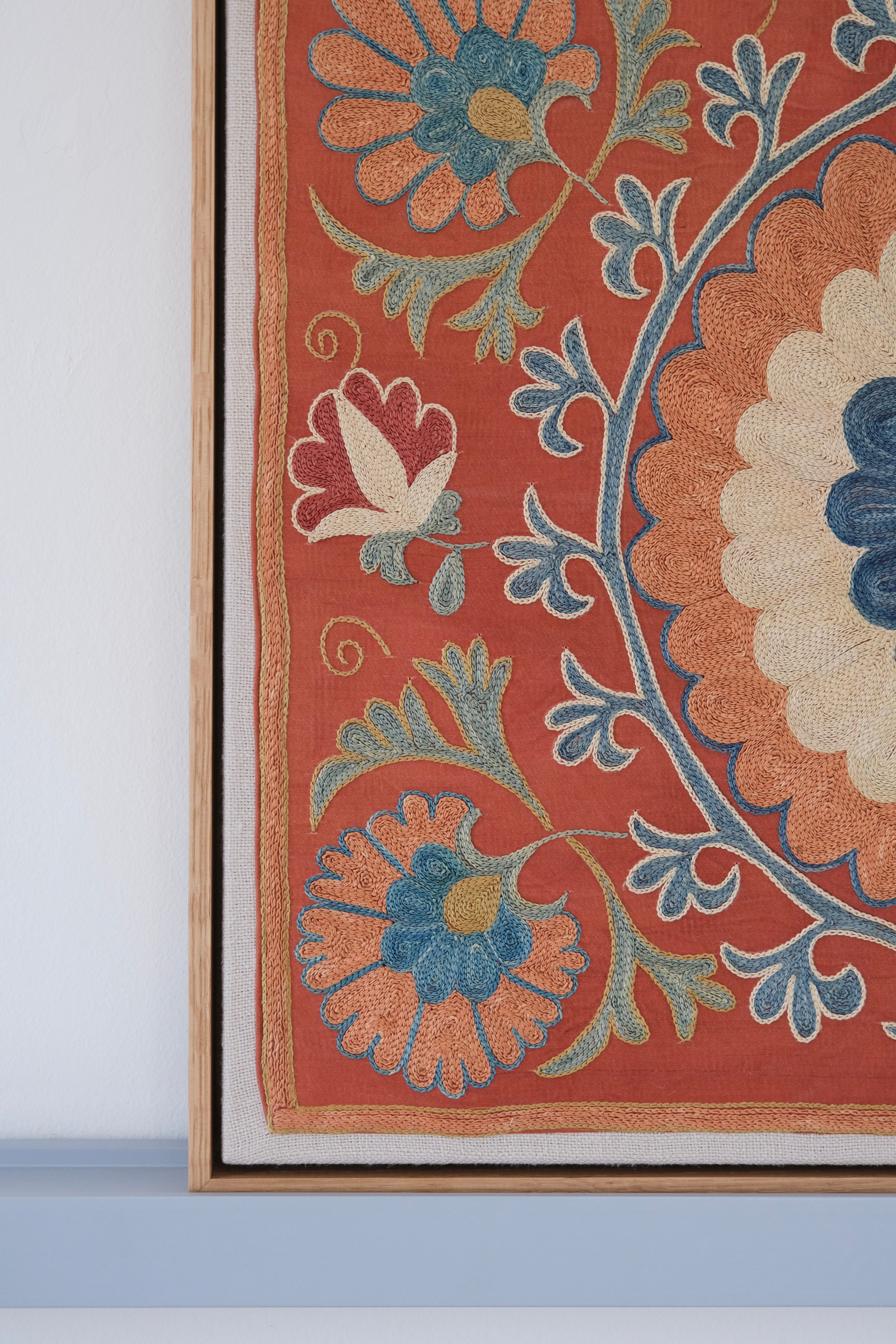 Framed suzani, embroidered textile art, wooden frame, dark orange with blue and green floral motifs, close-up