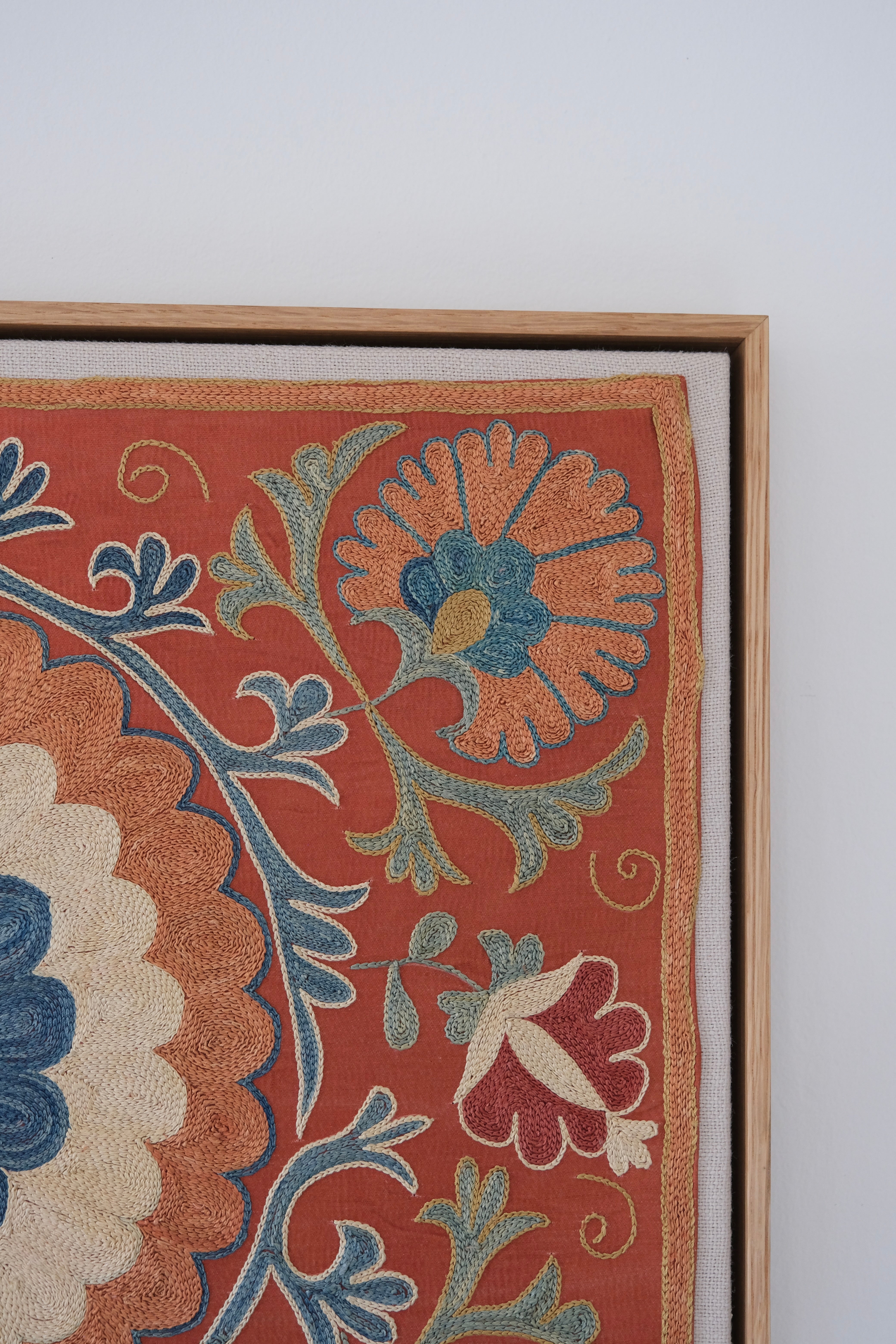 Framed suzani, embroidered textile art, wooden frame, dark orange with blue and green floral motifs, close-up
