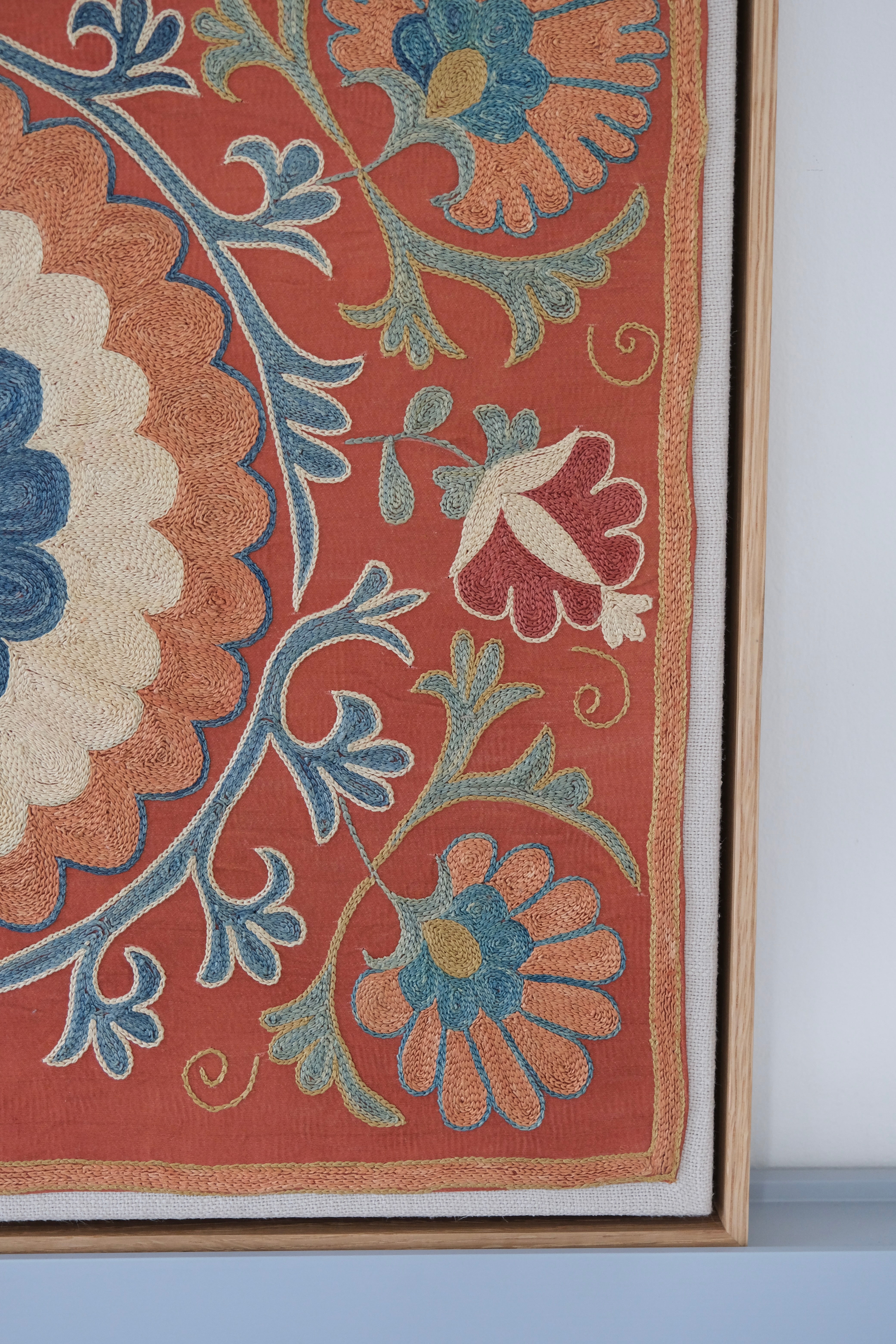 Framed suzani, embroidered textile art, wooden frame, dark orange with blue and green floral motifs, close-up